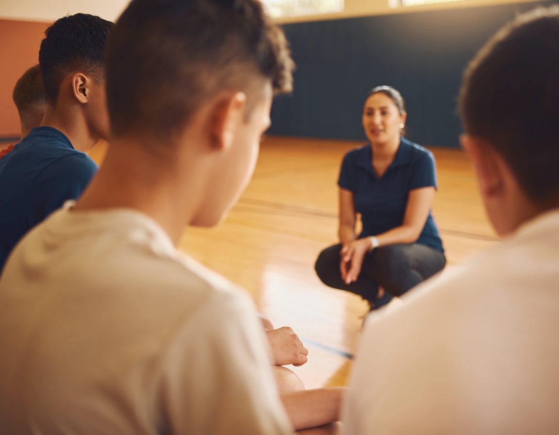 Coach crouching to talk with teenage players on a gym bench
