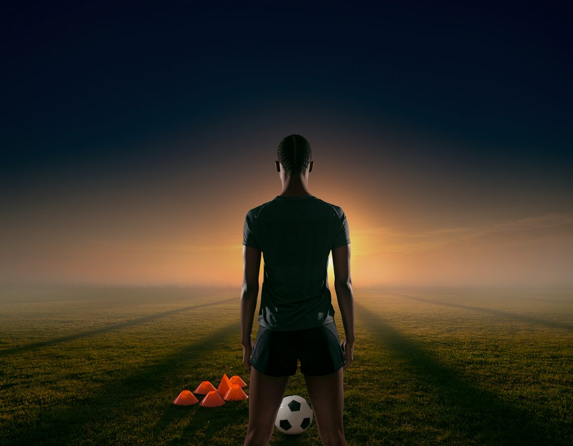 Soccer player standing on a misty training field at sunrise with a ball and cones